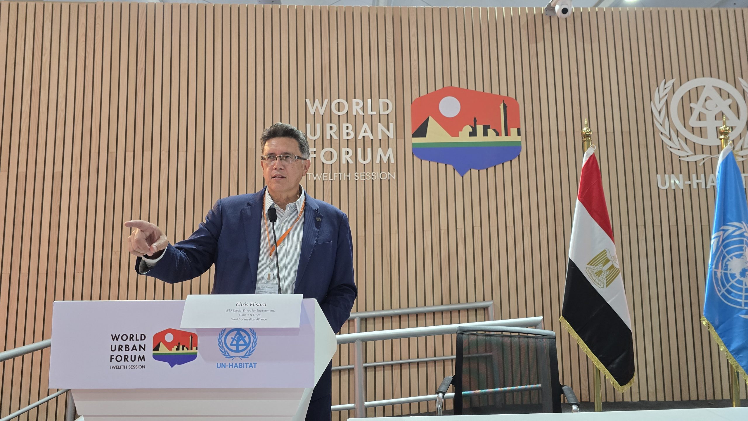 Dr. Chris Elisara speaking at a podium at the Faith Pavilion during the World Urban Forum (WUF12) in Cairo, with UN-Habitat and Egyptian flags behind him
