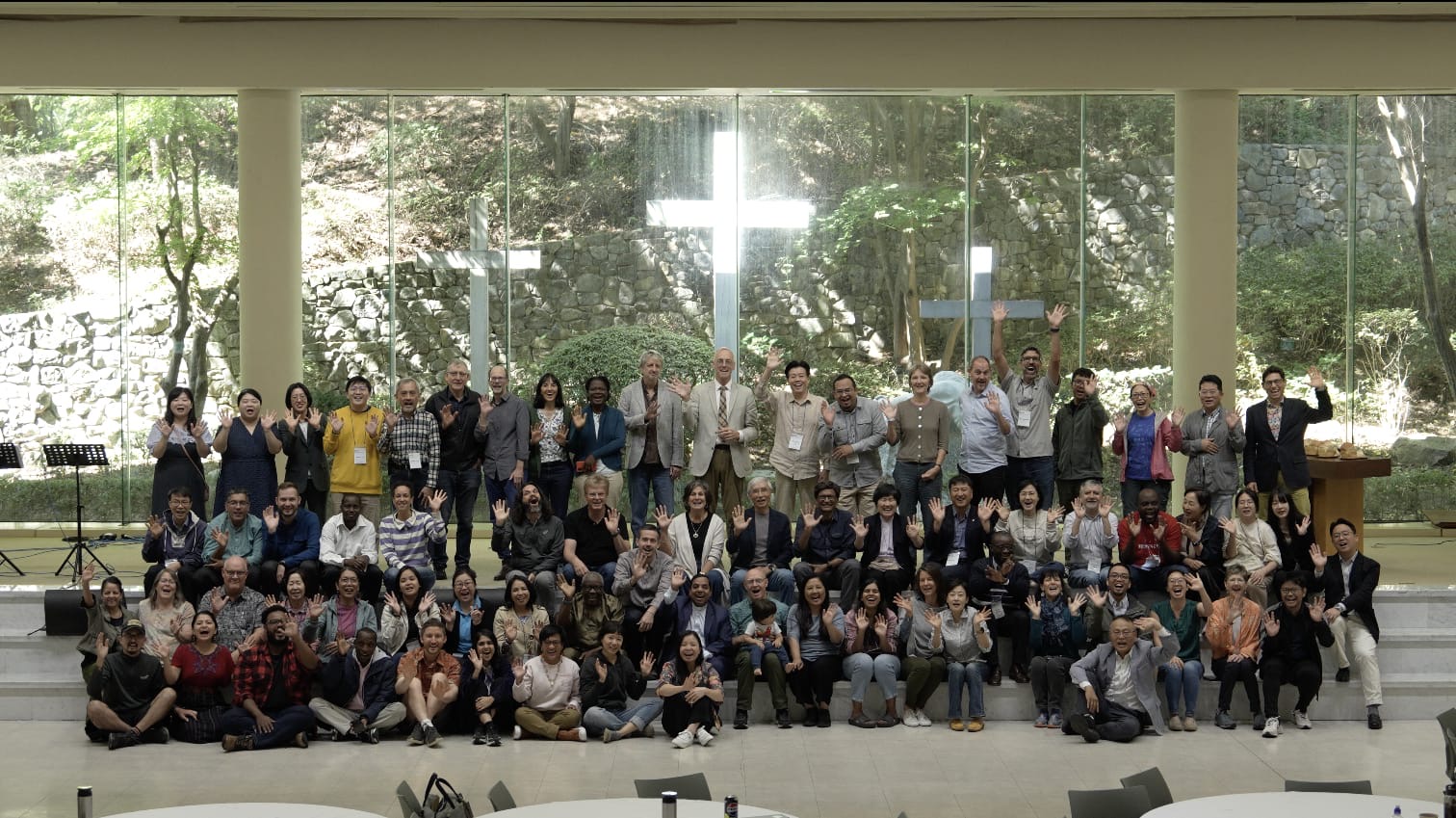 Large group photo of Global Creation Care Forum delegates gathered indoors at Somang Retreat Center in South Korea, waving with crosses visible through the windows behind them