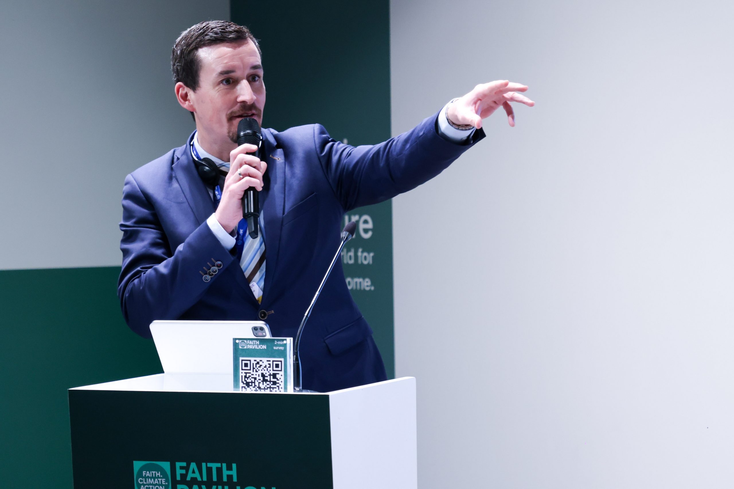 Matthias Boehning moderating the programme at the Faith Pavilion during COP28 in Dubai, standing at a podium with a Faith Pavilion QR code sign